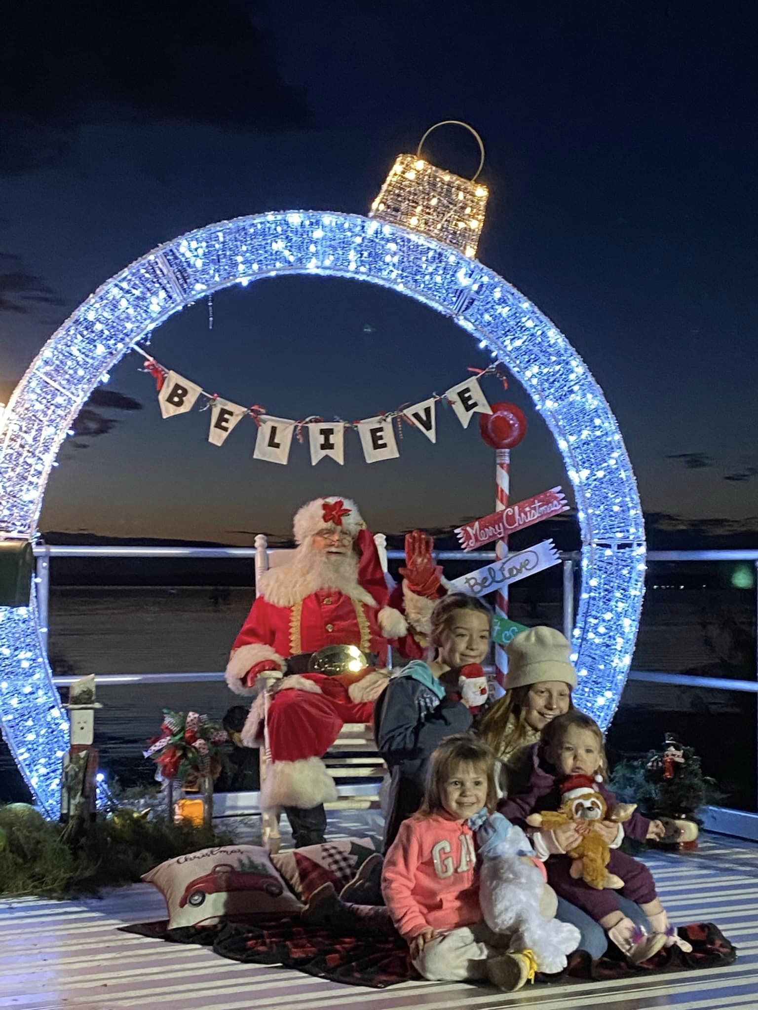 A photo of Santa Claus sitting on a bench, surrounded by children of various ages. They are all smiling and looking at the camera. The background shows a festive Christmas scene with decorated trees and lights.